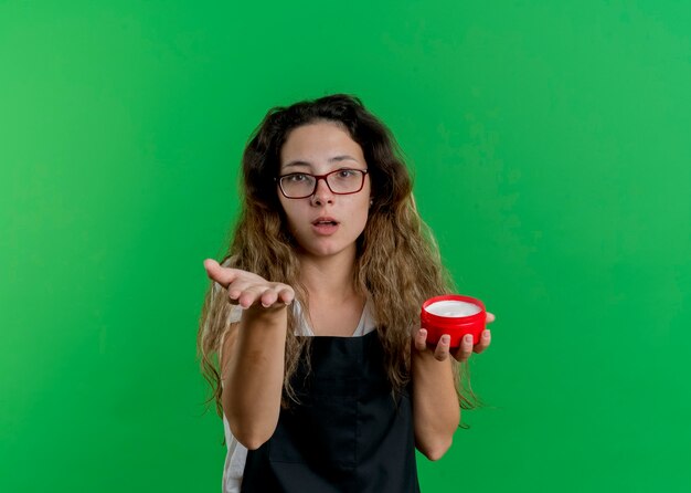 Mujer joven peluquero profesional en delantal sosteniendo un tarro de crema para el cabello mirando al frente con el brazo extendido como ofreciendo o pidiendo de pie sobre la pared verde