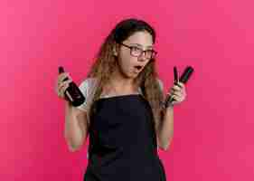 Foto gratuita mujer joven peluquero profesional en delantal con spray, cepillo de pelo y navaja mirando a un lado sorprendido y asombrado