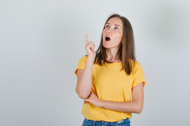 Mujer joven pensando mientras apunta el dedo hacia arriba en camiseta, pantalones cortos y mirando sorprendido