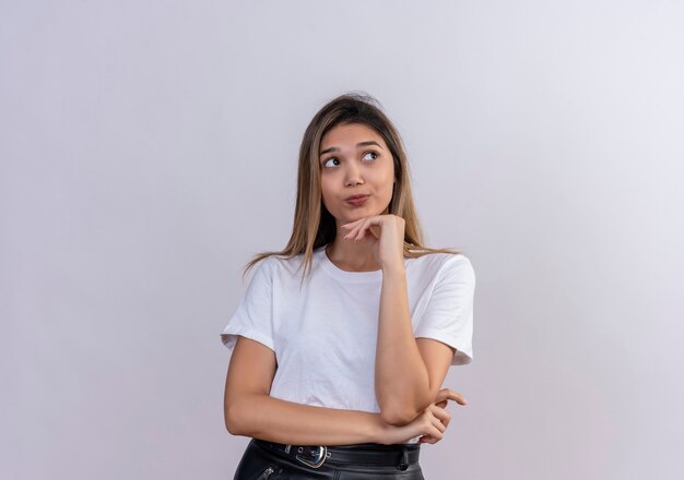 Una mujer joven pensante en camiseta blanca manteniendo la mano en la barbilla y mirando a otro lado mientras está de pie