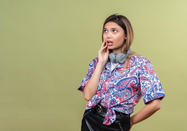 Una mujer joven pensativa con camisa estampada de paisley en auriculares pensando con las manos en la cara mientras mira hacia arriba