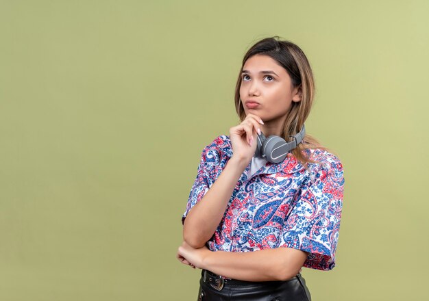 Una mujer joven pensativa en camisa estampada de paisley usando audífonos pensando mientras mira hacia arriba