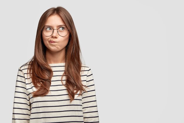 Mujer joven pensativa con gafas posando contra la pared blanca