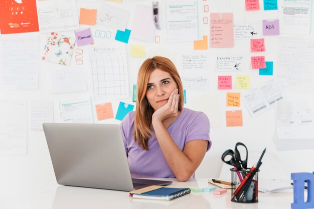 Mujer joven pensativa del pelirrojo que se sienta en el ordenador portátil contra la pared con las notas