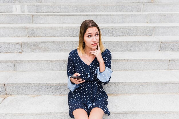 Mujer joven pensativa que se sienta en la escalera que sostiene el teléfono móvil