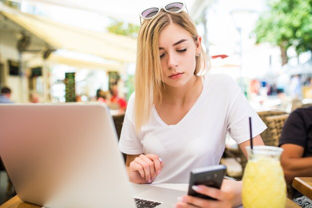 Mujer joven pensativa sostiene el teléfono, utiliza la computadora portátil para la comunicación en línea