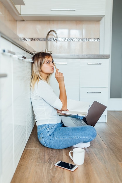 Mujer joven pensativa con taza de café y portátil en la cocina moderna y luminosa en casa.