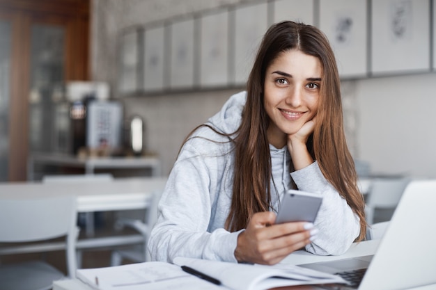 Mujer joven perfecta con teléfono inteligente y computadora portátil navegando a través de fotos mirando a la cámara sonriendo regocijo.