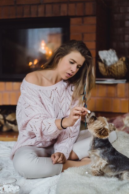 Mujer joven con un perro cerca de la chimenea bebe cacao con malvaviscos.