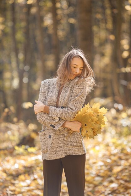 Mujer joven de pie en el bosque de otoño. Mujer morena sosteniendo hojas amarillas. Chica con chaqueta marrón de moda.