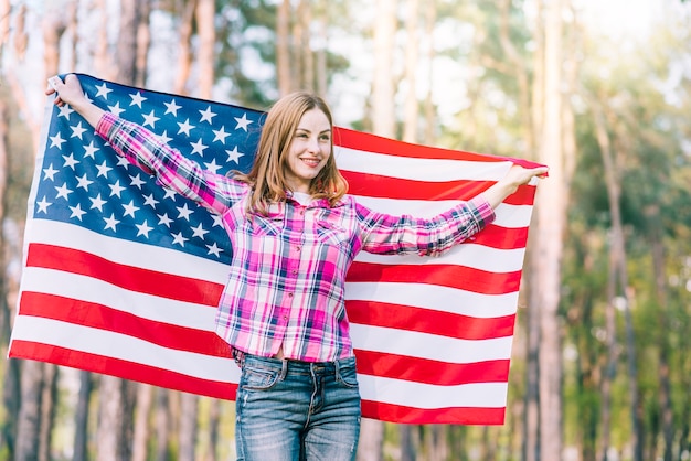 Mujer joven de pie en el bosque y sosteniendo la bandera de Estados Unidos