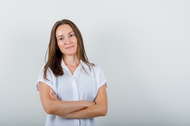 Mujer joven de pie con los brazos cruzados en blusa blanca y luciendo hermosa.