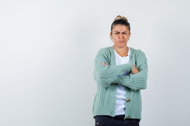 Mujer joven de pie con los brazos cruzados en camisa blanca y chaqueta de punto verde menta y mirando enojado