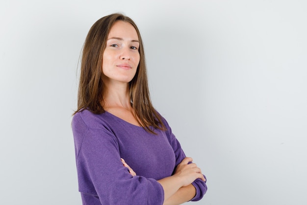 Mujer joven de pie con los brazos cruzados en camisa violeta y mirando confiado, vista frontal.