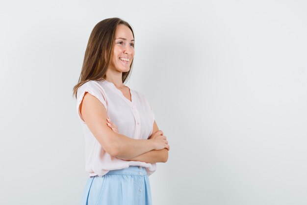 Mujer joven de pie con los brazos cruzados en camiseta, falda y mirando positivo, vista frontal.