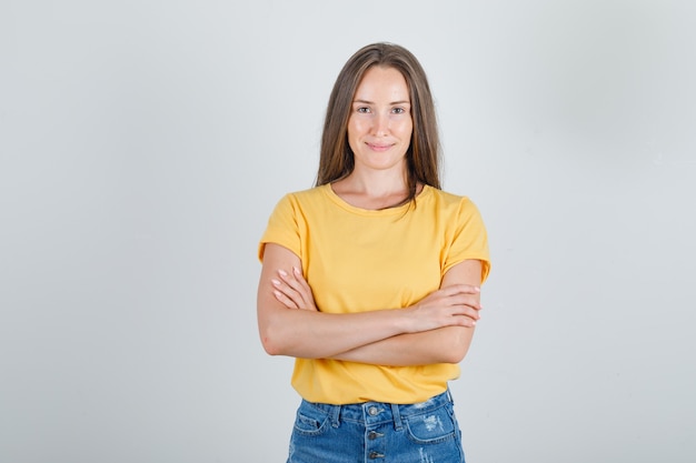 Mujer joven de pie con los brazos cruzados en camiseta, pantalones cortos y aspecto alegre.