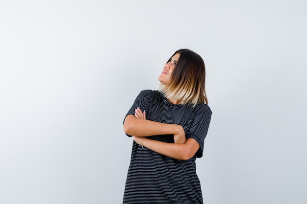 Foto gratuita mujer joven de pie con los brazos cruzados mientras mira hacia arriba con vestido de polo y mira pensativa, vista frontal.