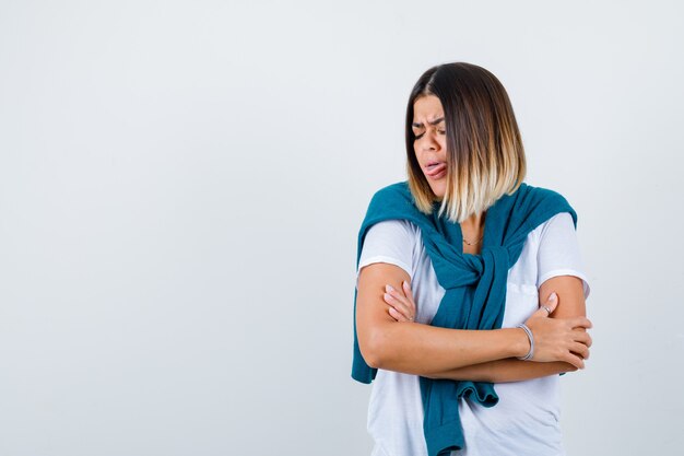 Mujer joven de pie con los brazos cruzados mientras saca la lengua en una camiseta blanca y parece disgustado, vista frontal.