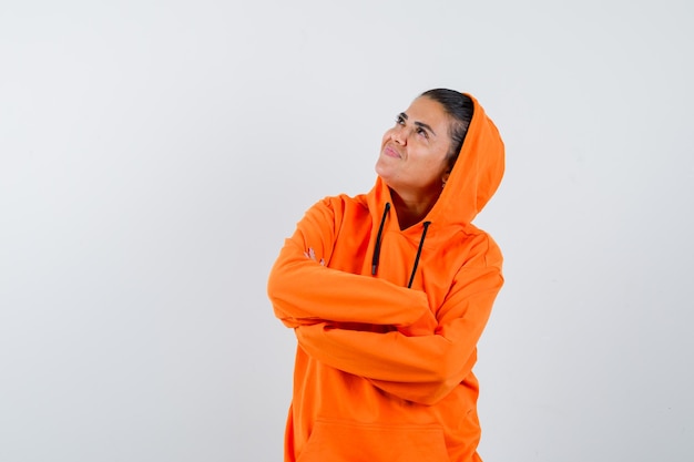 Mujer joven de pie con los brazos cruzados, mirando a otro lado en una sudadera con capucha naranja y luciendo radiante