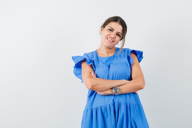 Mujer joven de pie con los brazos cruzados en vestido azul y mirando alegre