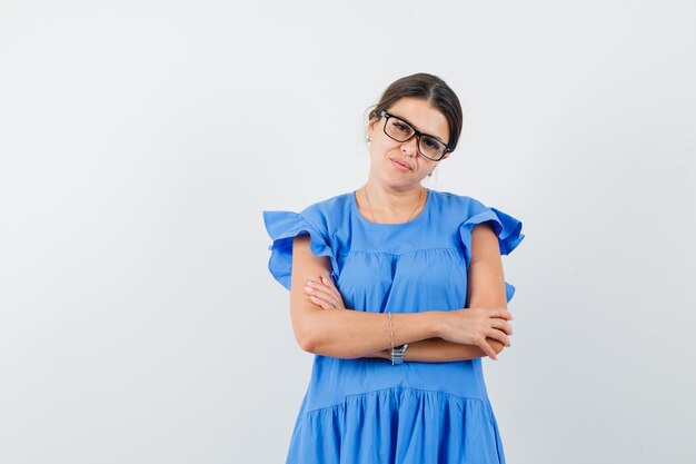 Mujer joven de pie con los brazos cruzados en vestido azul y mirando inteligente