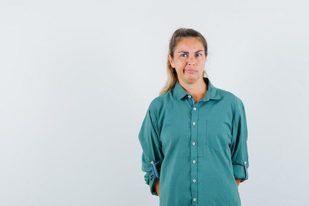 Mujer joven de pie con la espalda recta, haciendo muecas y posando al frente en blusa verde y mirando pensativo