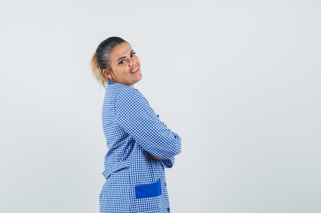 Mujer joven de pie con la espalda recta mientras mira por encima del hombro en camisa de pijama de cuadros vichy azul y mirando bonita, vista frontal.