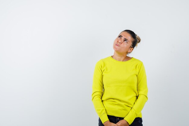 Mujer joven de pie con la espalda recta, mirando hacia arriba y posando a la cámara en suéter amarillo y pantalón negro y mirando pensativo