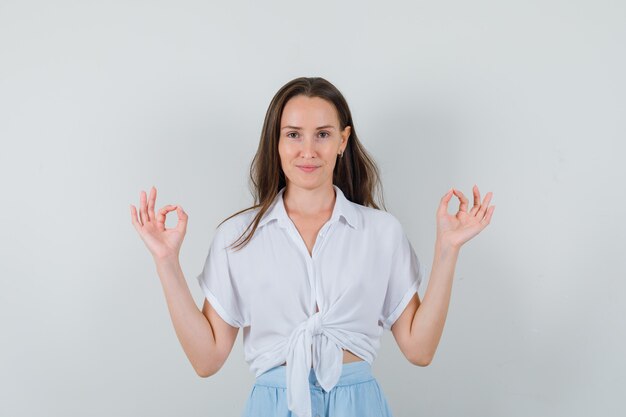 Mujer joven de pie con la espalda recta, mostrando el signo de ok con ambas manos en una blusa blanca y una falda azul claro y luciendo confiada