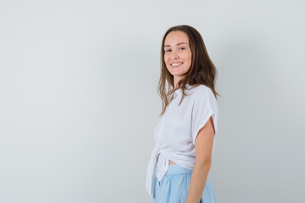 Mujer joven de pie con la espalda recta y posando al frente con blusa blanca y falda azul claro y mirando alegre