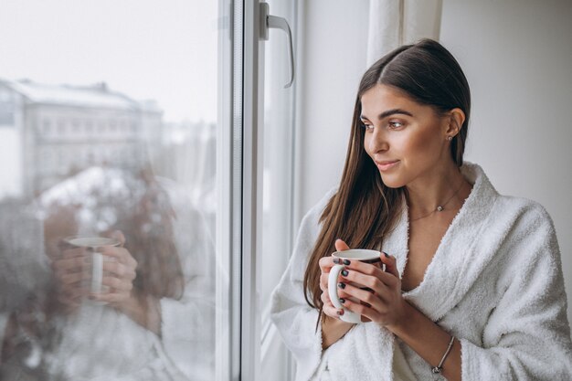 Mujer joven de pie junto a la ventana tomando café caliente