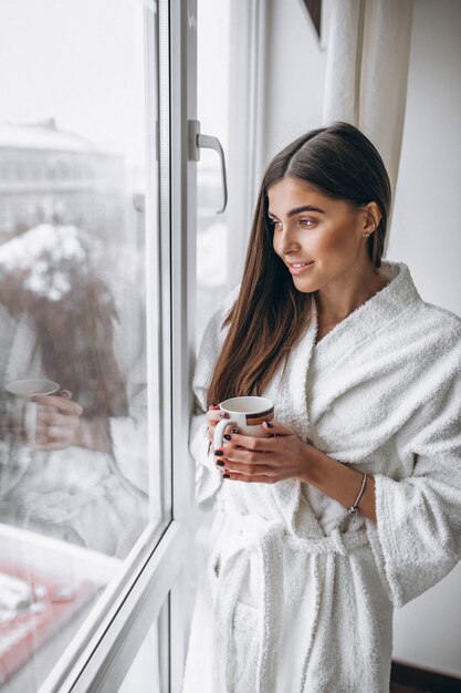 Mujer joven de pie junto a la ventana tomando café caliente