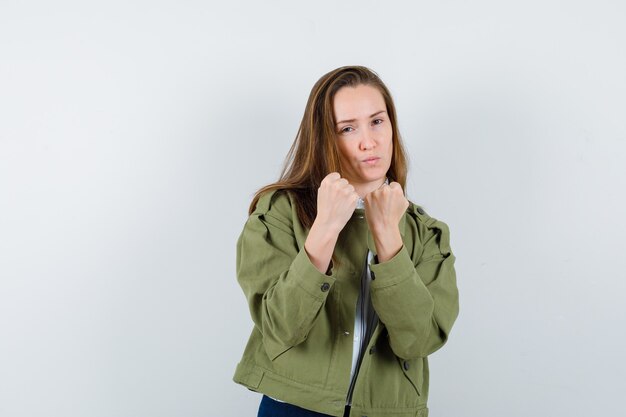 Mujer joven de pie en pose de lucha en camisa, chaqueta y mirando confiado. vista frontal.