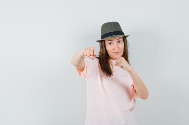 Mujer joven de pie en pose de lucha en camiseta rosa, sombrero y mirando confiado, vista frontal.