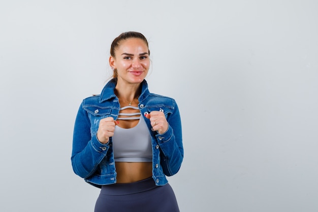 Mujer joven de pie en pose de lucha en top corto, chaqueta, pantalón y mirando confiado. vista frontal.