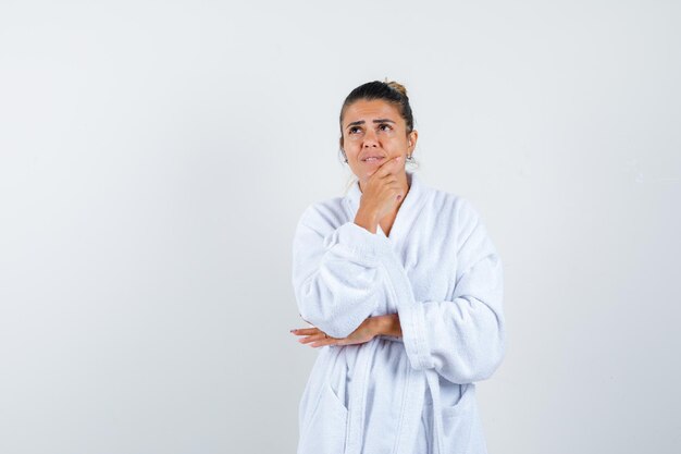Mujer joven de pie en pose de pensamiento en bata de baño y mirando pensativo