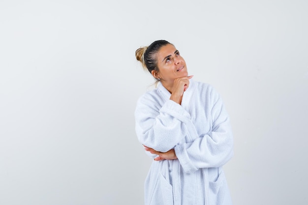 Mujer joven de pie en pose de pensamiento en bata de baño y mirando pensativo