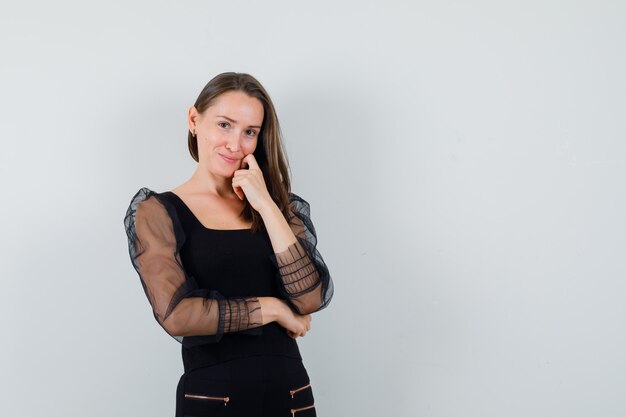 Mujer joven de pie en pose de pensamiento en blusa negra y pantalón negro y mirando pensativo, vista frontal.