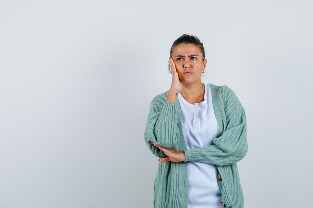 Mujer joven de pie en pose de pensamiento en camisa blanca y chaqueta de punto verde menta y mirando pensativo