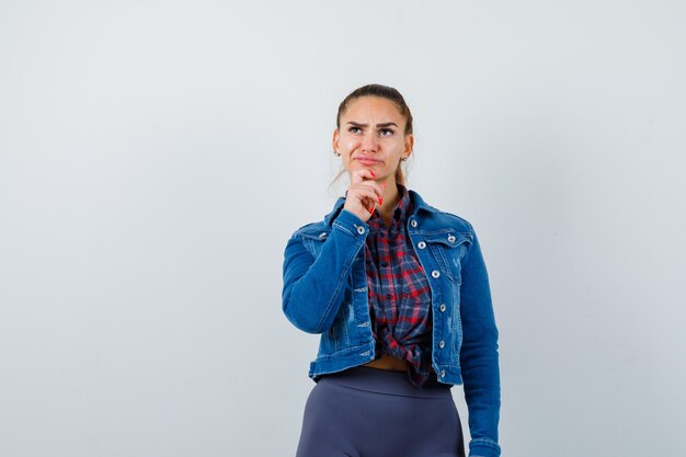 Mujer joven de pie en pose de pensamiento en camisa a cuadros, chaqueta, pantalón y mirando pensativo. vista frontal.