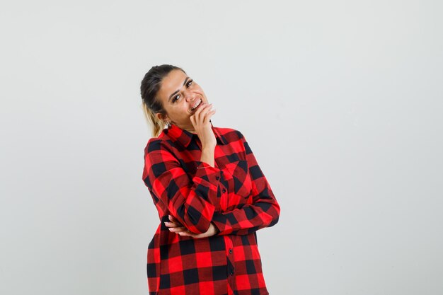 Mujer joven de pie en pose de pensamiento en camisa a cuadros y mirando alegre, vista frontal.