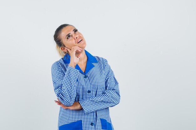 Mujer joven de pie en pose de pensamiento en camisa de pijama de cuadros vichy azul y mirando pensativo, vista frontal.