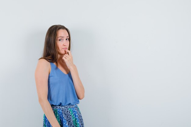 Mujer joven de pie en pose de pensamiento en camiseta, falda y mirada sensata. vista frontal.