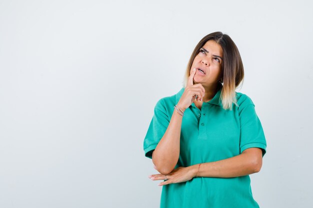 Mujer joven de pie en pose de pensamiento en camiseta de polo y mirando perplejo, vista frontal.