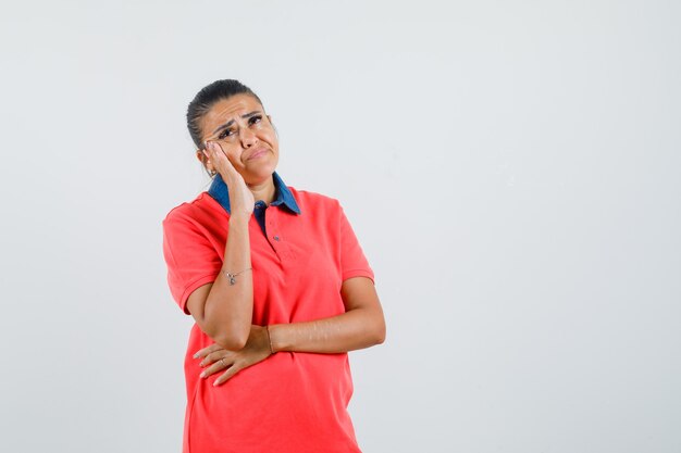 Mujer joven de pie en pose de pensamiento en camiseta roja y mirando pensativo, vista frontal.