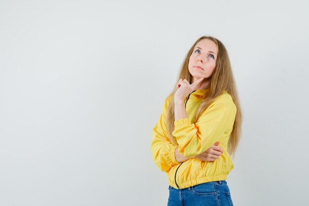 Mujer joven de pie en pose de pensamiento en chaqueta de bombardero amarilla y jean azul y mirando pensativo, vista frontal.