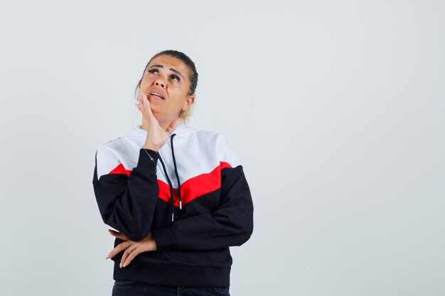 Mujer joven de pie en pose de pensamiento, cubriendo la boca con la mano en suéter y jeans negros y mirando pensativo. vista frontal.