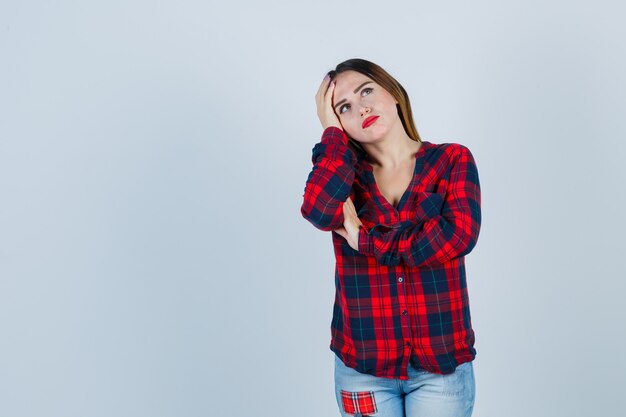 Mujer joven de pie en pose de pensamiento, con la mano en la cabeza en camisa a cuadros, jeans y mirando pensativo. vista frontal.