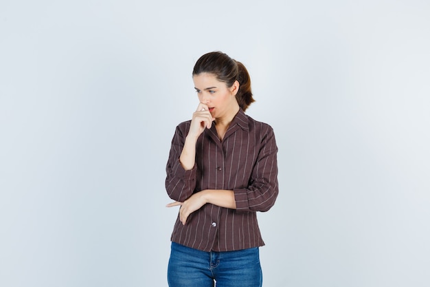 Mujer joven de pie en pose de pensamiento, con la mano delante de la boca en camisa a rayas, jeans y mirando pensativo, vista frontal.