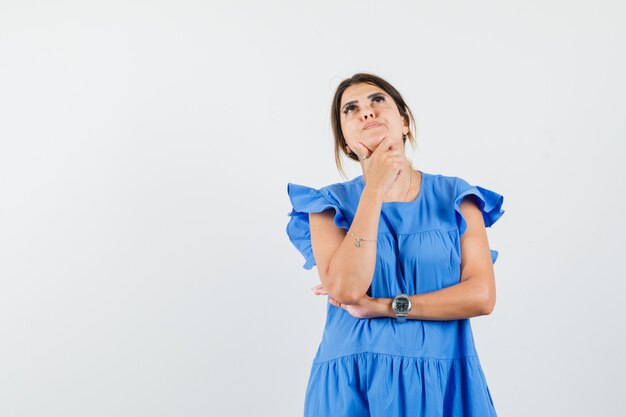 Mujer joven de pie en pose de pensamiento mientras mira hacia arriba con vestido azul y parece esperanzado